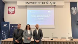 Trzy osoby stoją przed stołem konferencyjnym w sali Uniwersytetu Śląskiego; w tle widoczny jest ekran z prezentacją dotyczącą konsultacji projektu Strategii Rozwoju Szkolnictwa Wyższego oraz godło Polski i symbole uczelni.