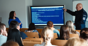 Policjant prowadzi prezentację przed grupą słuchaczy w sali wykładowej, wskazując na slajd wyświetlany na ekranie, obok stojącej prowadzącej.