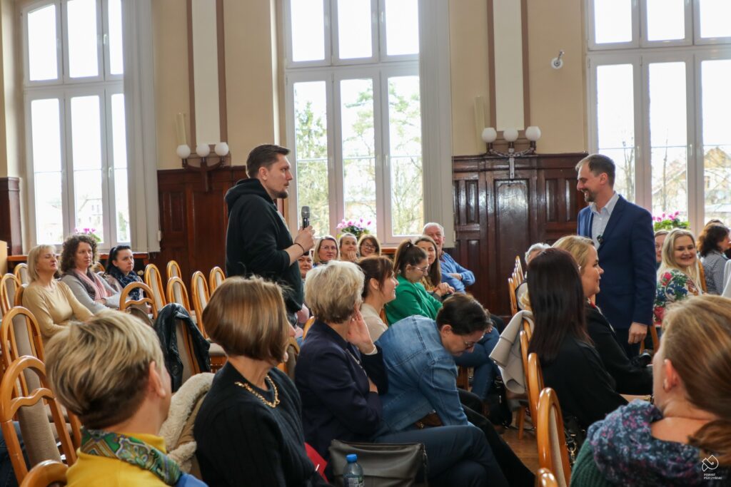 Uczestnik spotkania stoi i z mikrofonem w ręku zadaje pytanie prowadzącemu wykład, natomiast zgromadzeni w sali słuchacze uważnie obserwują rozmowę.