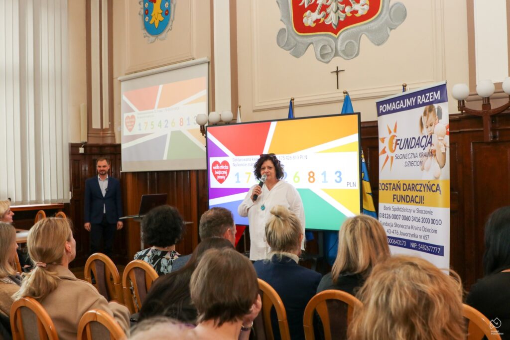 Kobieta przemawia przez mikrofon  na tle ekranu z liczbą zebranych środków, a mężczyzna stoi obok.