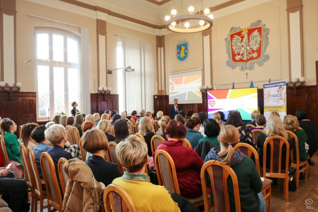 Widok na salę wykładową z udziałem uczestników wydarzenia, z prowadzącym prezentację na tle ekranu z liczbą zebranych funduszy.
