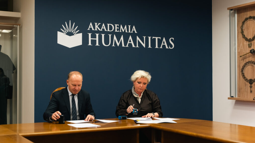 Dwie osoby siedzą przy stole i podpisują dokumenty; w tle granatowa ściana z logo Akademii Humanitas.
