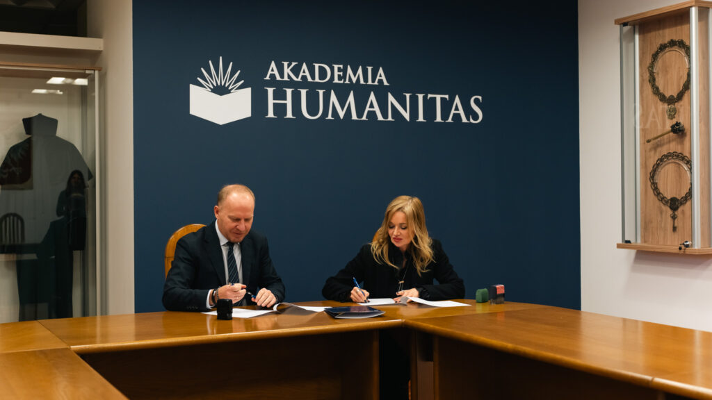 Dwie osoby siedzą przy stole i podpisują dokumenty; w tle granatowa ściana z logo Akademii Humanitas.