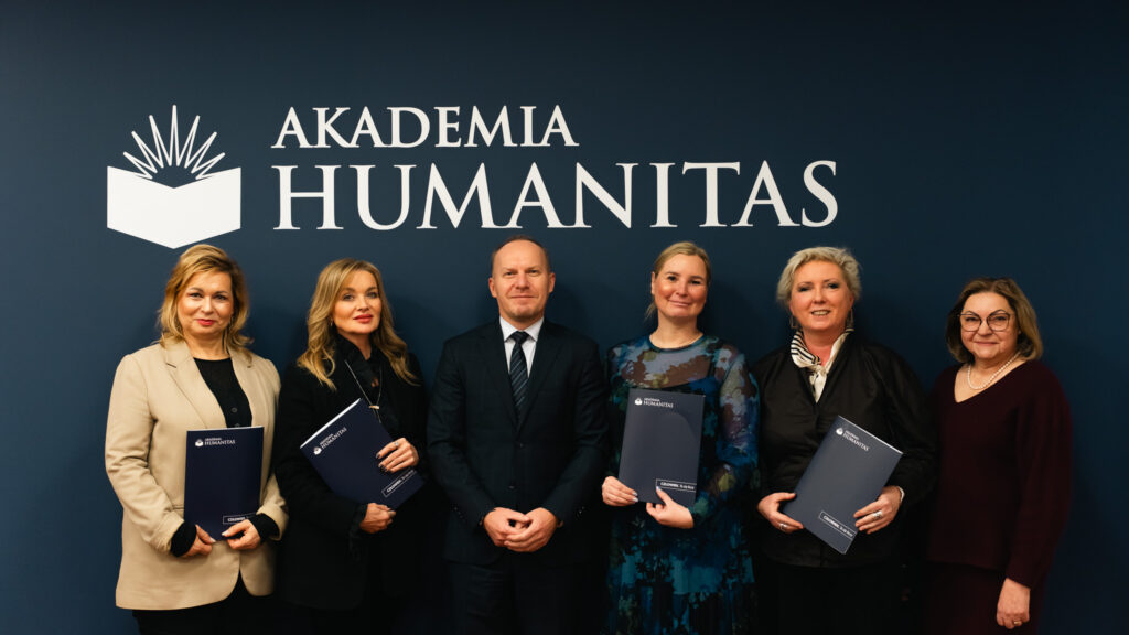 Grupowe zdjęcie sześciu osób stojących przed ścianką z logo Akademii Humanitas; uczestnicy trzymają teczki z logo uczelni, w centrum mężczyzna w garniturze.