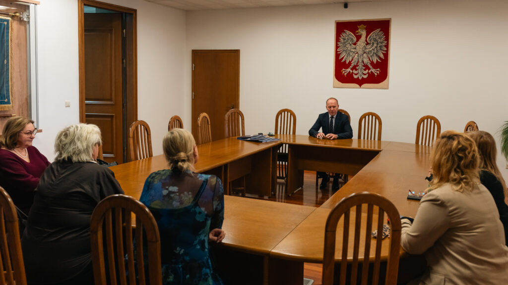 Spotkanie w sali konferencyjnej przy dużym stole; kilka osób siedzi naprzeciw mężczyzny w garniturze, na ścianie widoczny godło Polski.