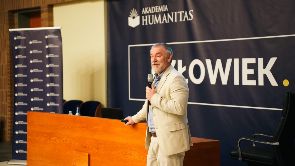Prelegent w jasnym garniturze przemawia z mikrofonem podczas wykładu; w tle baner Akademii Humanitas z hasłem »Człowiek. To się liczy«.