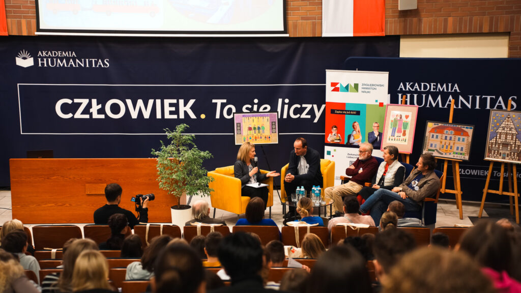 Panel dyskusyjny w auli Akademii Humanitas: prowadząca z mikrofonem rozmawia z zaproszonymi gośćmi siedzącymi na fotelach, w tle baner «Człowiek. To się liczy», logo Zagłębiowskiego Maratonu Nauki oraz wystawione prace plastyczne, na widowni zgromadzona publiczność.