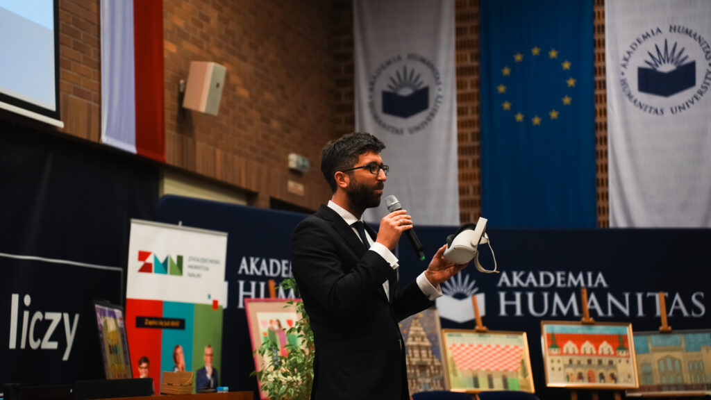 Mężczyzna w garniturze przemawia do publiczności, trzymając mikrofon i prezentując gogle wirtualnej rzeczywistości (VR); w tle banery Akademii Humanitas, flaga Unii Europejskiej oraz wystawione obrazy.