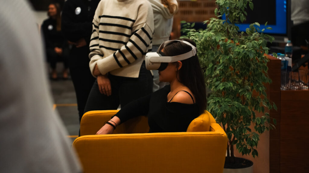 Kobieta siedząca w żółtym fotelu korzysta z gogli wirtualnej rzeczywistości (VR) podczas wydarzenia; w tle stoją inni uczestnicy oraz roślina doniczkowa.