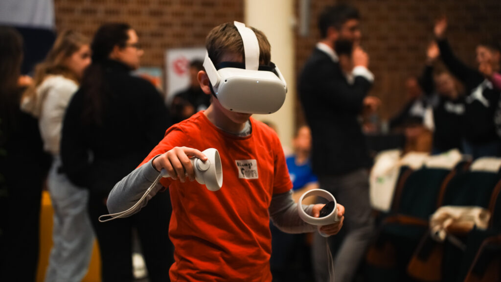 Chłopiec w czerwonej koszulce korzysta z gogli wirtualnej rzeczywistości (VR), trzymając kontrolery w dłoniach, podczas wydarzenia edukacyjnego; w tle widoczni są inni uczestnicy.