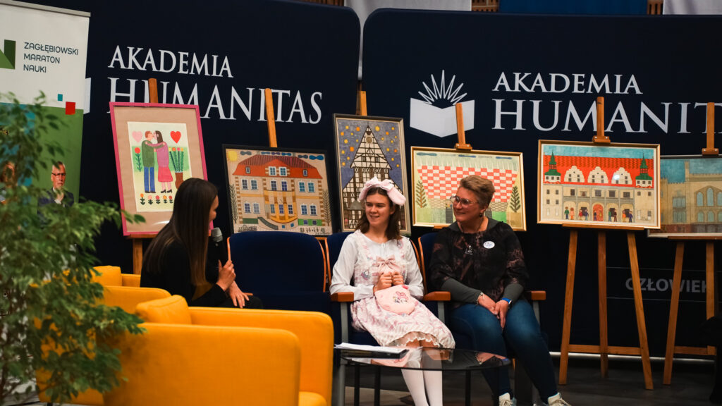 Rozmowa na scenie podczas wydarzenia w Akademii Humanitas: prowadząca z mikrofonem rozmawia z dwiema siedzącymi uczestniczkami. W tle stoją sztalugi z kolorowymi ilustracjami budynków i postaci oraz banery Akademii Humanitas.