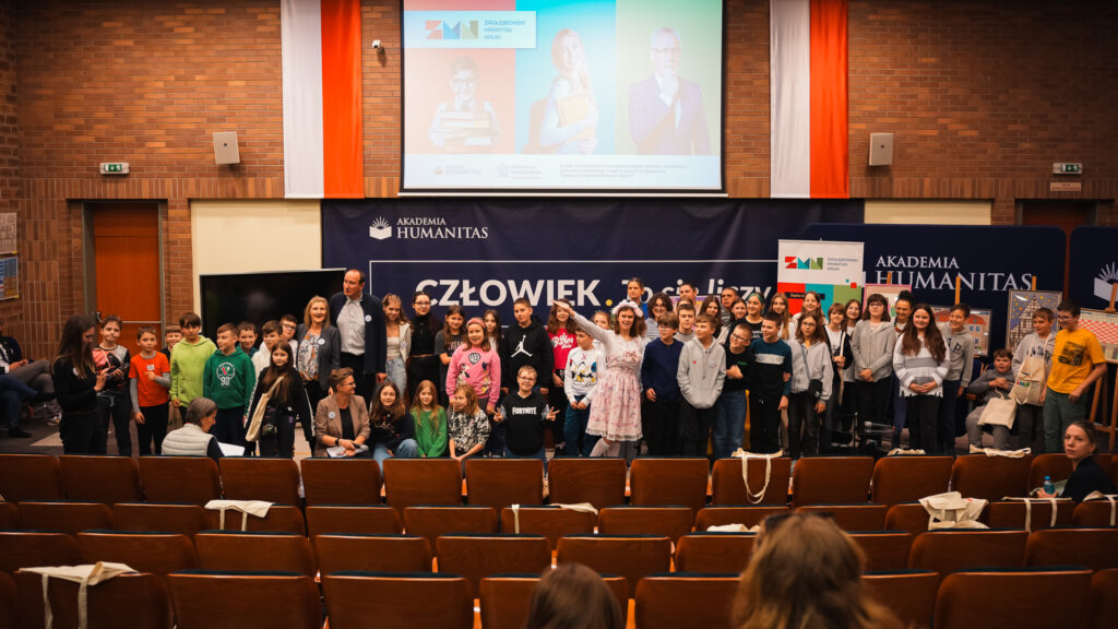 Grupowe zdjęcie dzieci, młodzieży i dorosłych uczestników wydarzenia edukacyjnego stojących na scenie auli, na tle banerów Akademii Humanitas i hasła „Człowiek. To się liczy”, po zakończeniu spotkania.