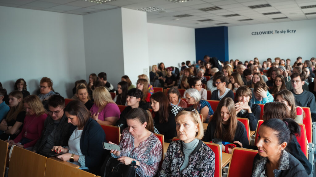 Publiczność siedząca w sali wykładowej na czerwonych krzesłach, złożona z młodzieży i dorosłych; uczestnicy wydarzenia edukacyjnego przeglądają telefony, rozmawiają i czekają na rozpoczęcie wykładu. Na ścianie widoczny napis: »Człowiek. To się liczy«.