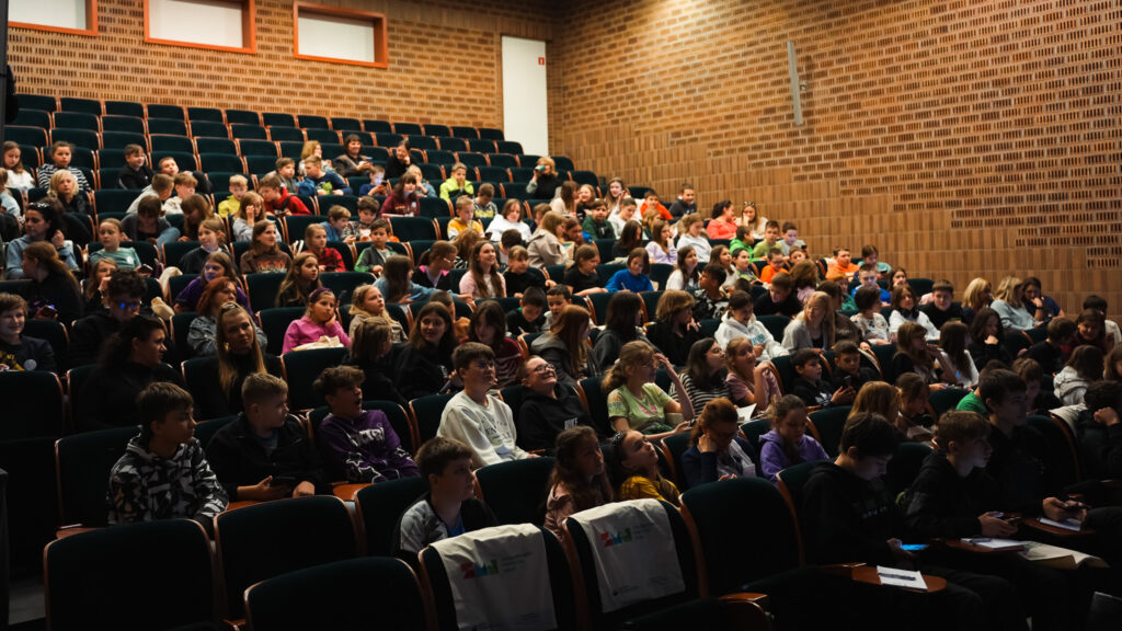 Widownia w auli wypełniona dziećmi i młodzieżą siedzącymi na zielonych fotelach, uczestniczącymi w wydarzeniu edukacyjnym; uczniowie rozmawiają i czekają na rozpoczęcie zajęć.