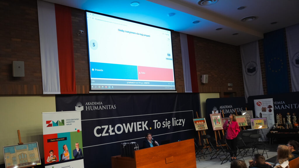 Prezentacja multimedialna wyświetlana na dużym ekranie w auli Akademii Humanitas, z pytaniem quizowym dotyczącym autyzmu; na scenie prowadzące wydarzenie, banery instytucji oraz elementy wystawy, a na widowni uczestnicy spotkania edukacyjnego.
