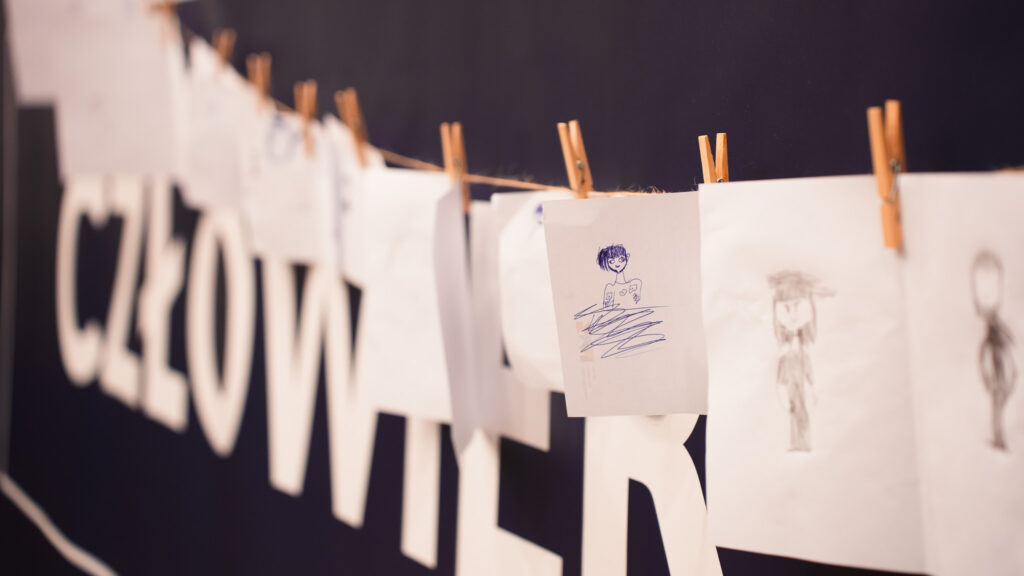 Rysunki dzieci zawieszone na sznurku za pomocą drewnianych klamerek, prezentowane na tle napisu scenicznego podczas wydarzenia edukacyjnego.