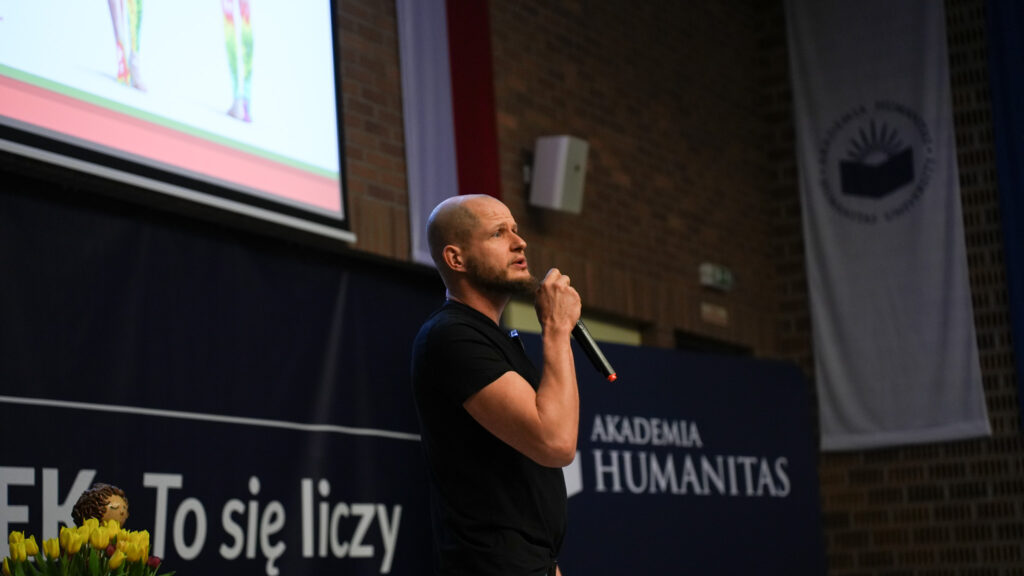 Prelegent z mikrofonem prowadzi wykład na scenie; w tle ekran z prezentacją i baner Akademii Humanitas.