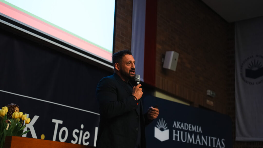 Prelegent z mikrofonem wygłasza wystąpienie na scenie; w tle baner Akademii Humanitas.