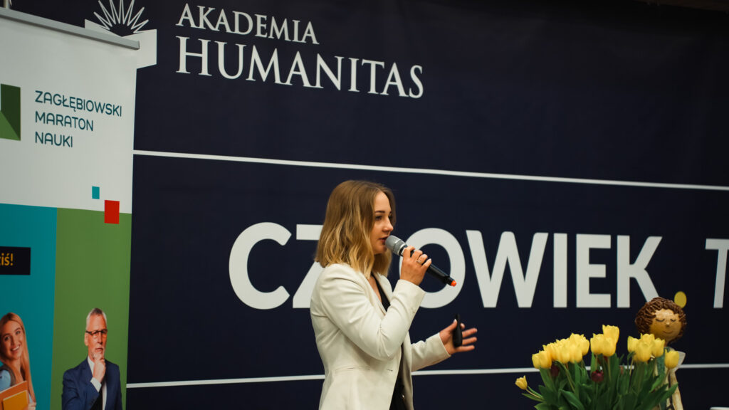 Prelegentka z mikrofonem prowadzi wystąpienie na scenie; w tle baner Akademii Humanitas.