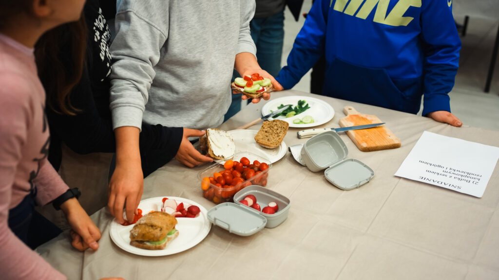 Osoby wspólnie przygotowują zdrowe kanapki przy stole, układając warzywa i pieczywo na talerzach.
