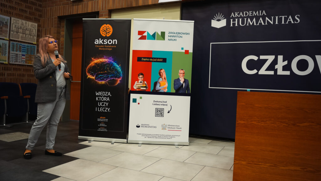 Prelegentka z mikrofonem prowadzi wykład; obok roll-upy partnerów i baner wydarzenia.