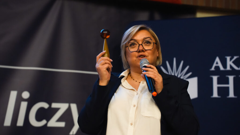 Prowadząca z mikrofonem prezentuje przedmiot podczas wykładu na scenie.