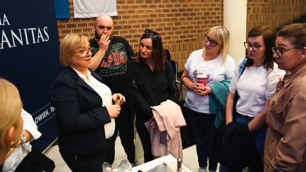 Grupa dorosłych uczestników słucha prowadzącej, która omawia temat podczas spotkania; rozmowa odbywa się przy stoisku wydarzenia.