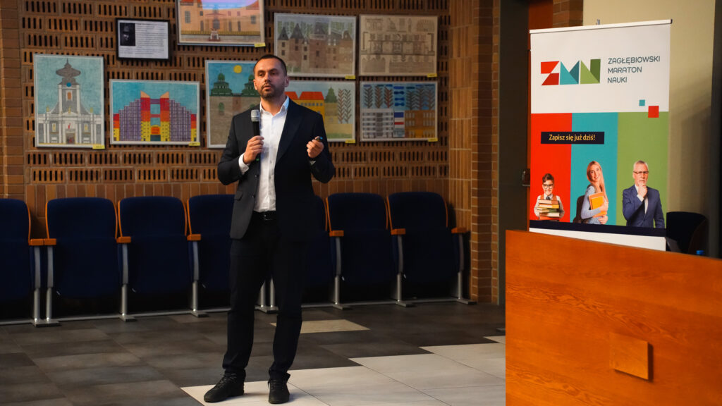 Prelegent w garniturze przemawia do publiczności, trzymając mikrofon; obok widoczny roll-up „Zagłębiowski Maraton Nauki”.