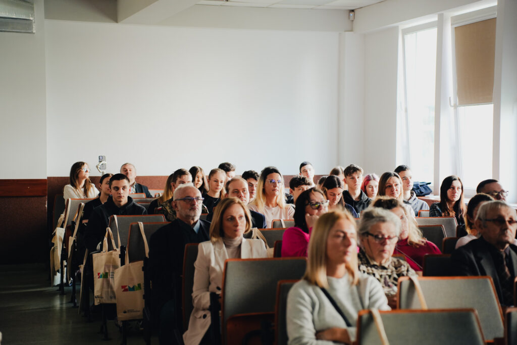 Publiczność siedząca w sali wykładowej, ujęcie od przodu. W kilku rzędach widoczne są osoby w różnym wieku, głównie młodzież i dorośli, skupieni na wydarzeniu odbywającym się poza kadrem. Uczestnicy siedzą na tapicerowanych krzesłach, część ma przy sobie torby materiałowe.