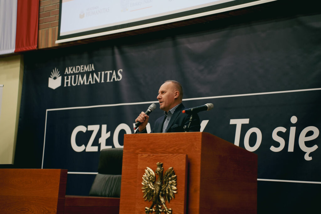 Mężczyzna w garniturze stoi przy mównicy z godłem orła i przemawia do publiczności, trzymając mikrofon w dłoni. Za nim widoczny jest duży baner z napisem „Akademia Humanitas”. Scena znajduje się w auli wykładowej.