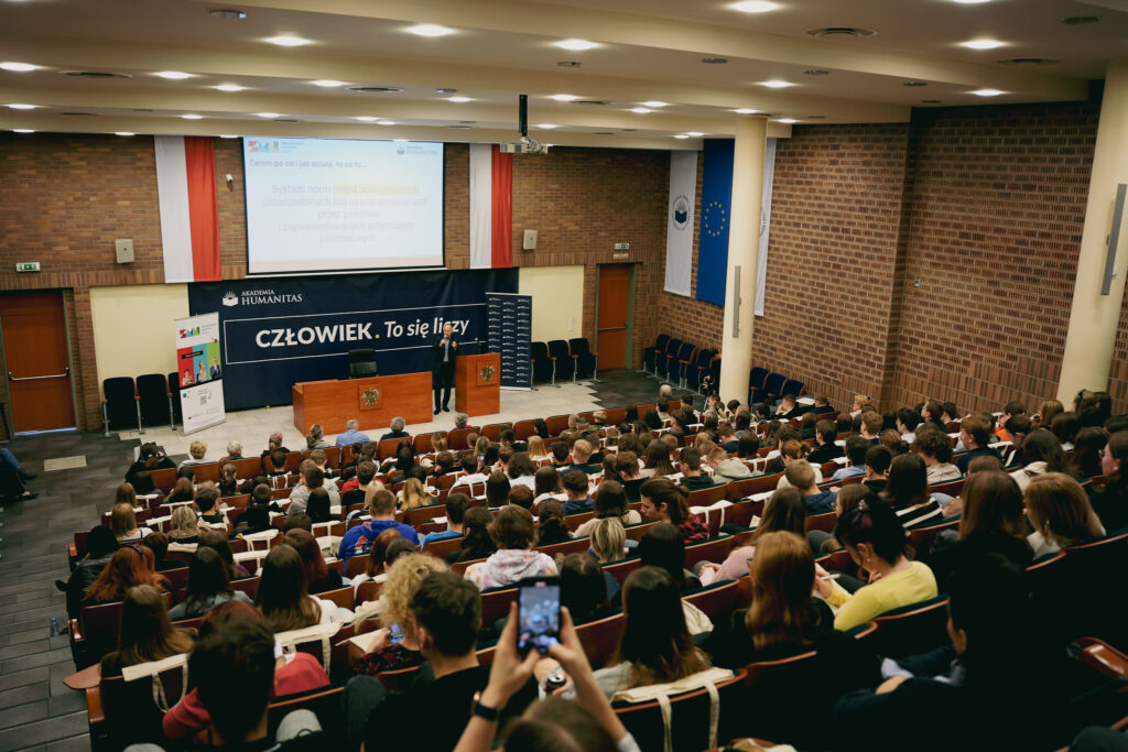 Szerokie ujęcie auli wypełnionej publicznością podczas wykładu. Na scenie, przy mównicy, stoi prelegent przemawiający do licznie zgromadzonych uczestników. Nad sceną widoczny jest ekran z prezentacją oraz baner z hasłem „Człowiek. To się liczy” i logo Akademii Humanitas.
