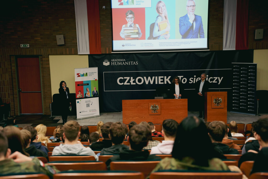 Sala wykładowa Akademii Humanitas podczas wydarzenia edukacyjnego – prelegenci stoją przy mównicach na scenie, a na ekranie wyświetlona jest prezentacja; na widowni siedzi licznie zgromadzona publiczność.