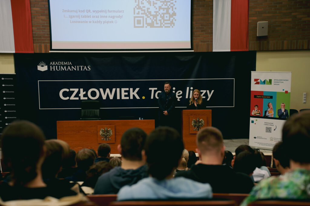 Widok na scenę w auli podczas wydarzenia akademickiego. Przy mównicy z godłem orła stoi kobieta i przemawia do publiczności; obok niej stoi mężczyzna. Za nimi znajduje się duży baner z napisem „Człowiek. To się liczy” oraz logo Akademii Humanitas. Nad sceną wyświetlany jest slajd z kodem QR i informacją o konkursie. Po prawej stronie widać roll-up „Zagłębiowski Maraton Nauki”. Na pierwszym planie widoczne są sylwetki siedzących uczestników.”