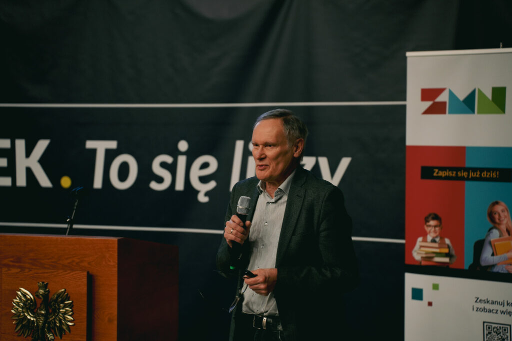 Prelegent przemawia do publiczności, trzymając mikrofon, na tle baneru „Człowiek. To się liczy” i roll-upu wydarzenia.