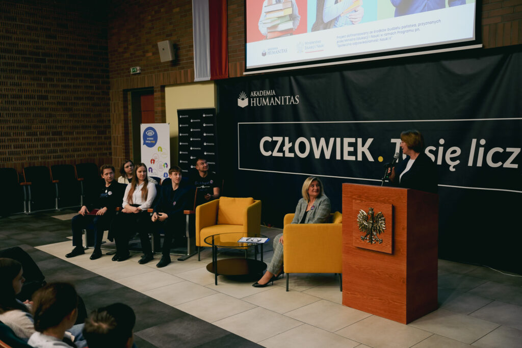 Panel dyskusyjny w auli: prowadząca przy mównicy mówi do mikrofonu, a obok siedzą uczestnicy panelu. W tle baner „Człowiek. To się liczy”