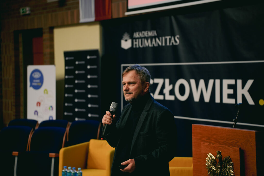 Mężczyzna ubrany w ciemny marynarkę i golf, stoi na scenie i przemawia do publiczności, trzymając mikrofon w dłoni. W tle widoczny jest baner z logo Akademii Humanitas i hasłem „Człowiek”, a obok fragment mównicy z godłem orła. 