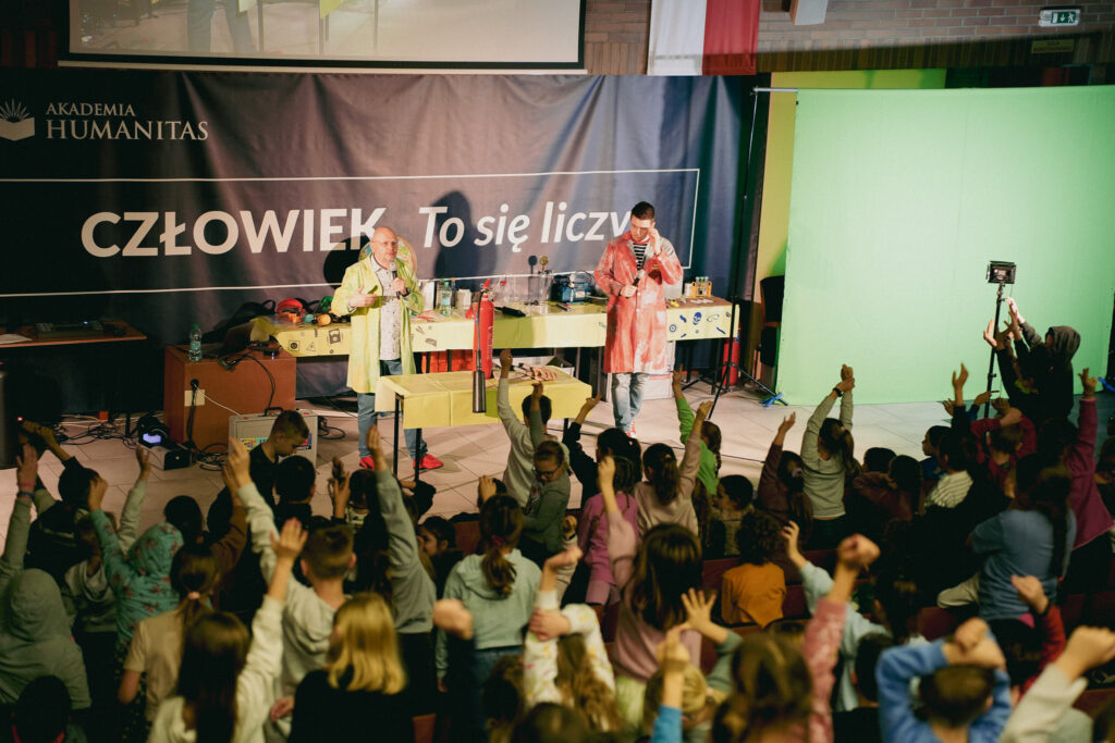 Pokaz naukowy na scenie: dwaj prowadzący w fartuchach wykonują eksperymenty, a dzieci na widowni podnoszą ręce i reagują entuzjastycznie.