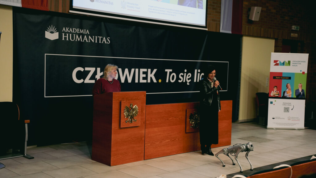 Dwie prelegentki na scenie auli; jedna przemawia do mikrofonu, obok mównica z godłem, a na podłodze stoi robot-pies. W tle baner „Człowiek. To się liczy”.