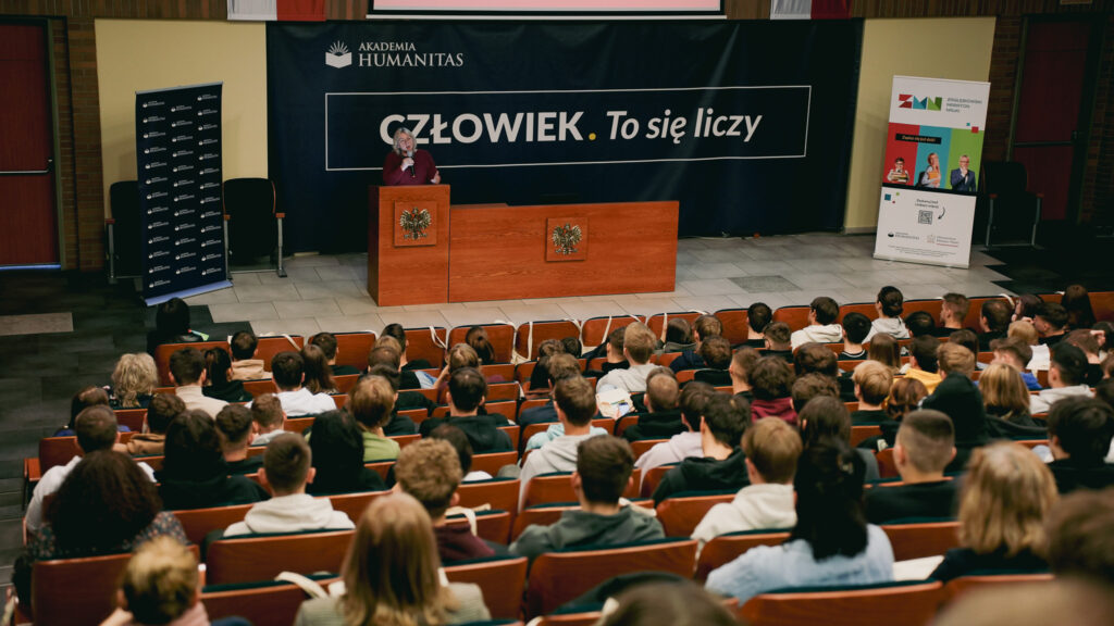 Prelegentka przemawia z mównicy do pełnej auli; w tle baner „Człowiek. To się liczy” Akademii Humanitas.