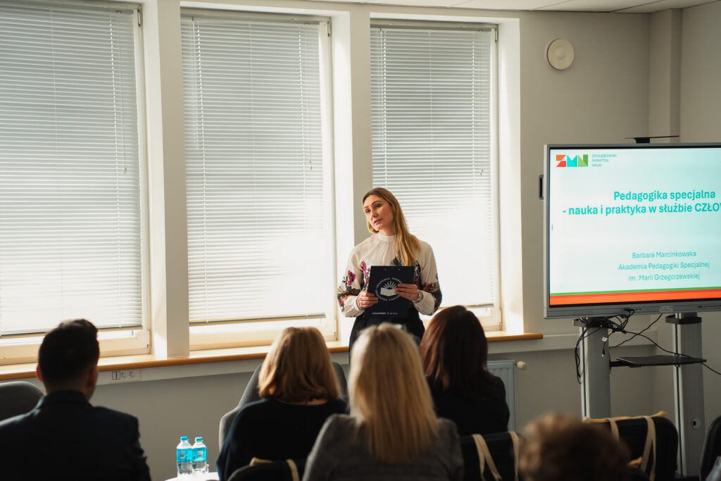 Prelegentka prowadzi wykład dla uczestników, stojąc obok ekranu z prezentacją.