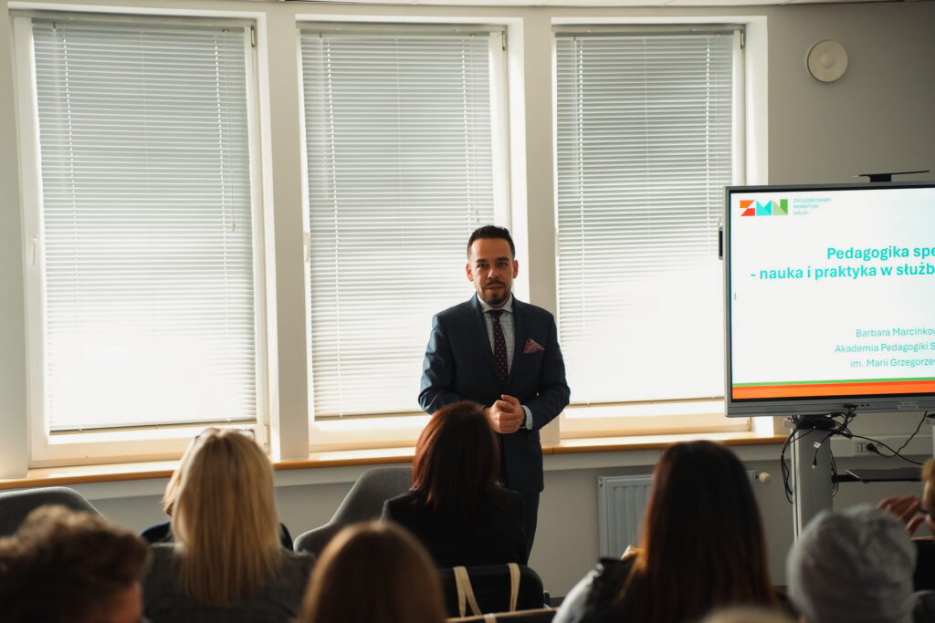 Prelegent w garniturze prowadzi wykład dla uczestników, stojąc obok ekranu z prezentacją.