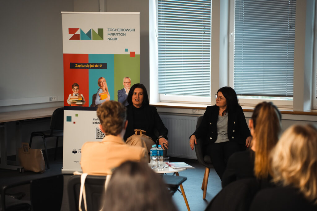 Dwie prowadzące siedzą i rozmawiają z uczestnikami podczas spotkania; w tle roll-up „Zagłębiowski Maraton Nauki”.