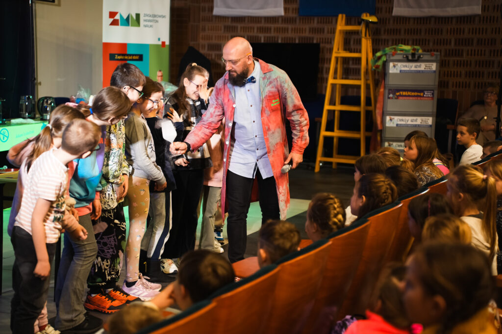 Prowadzący pokaz naukowy w kolorowym fartuchu angażuje dzieci stojące przy scenie, a pozostałe dzieci obserwują z widowni.