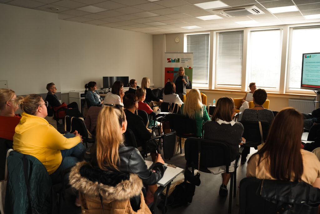 Warsztat w sali szkoleniowej: prowadząca rozmawia z uczestnikami siedzącymi w kręgu; w tle roll-up „Zagłębiowski Maraton Nauki”.