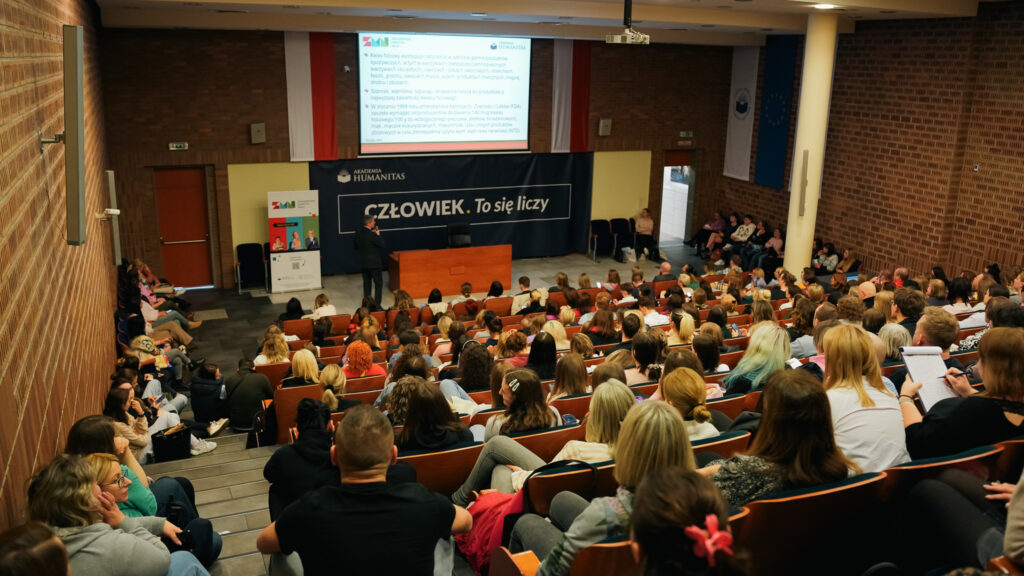 Widok z góry na aulę wypełnioną uczestnikami wykładu; na scenie prelegent prowadzi prezentację multimedialną, a za nim widoczny jest baner Akademii Humanitas z hasłem „Człowiek. To się liczy”.