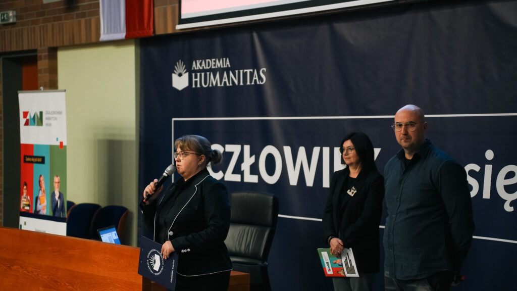 Kobieta przemawia do mikrofonu podczas wydarzenia w Akademii Humanitas; obok niej stoją dwie osoby, a w tle widoczny jest baner z hasłem »Człowiek. To się liczy« oraz roll-up Zagłębiowskiego Maratonu Nauki.