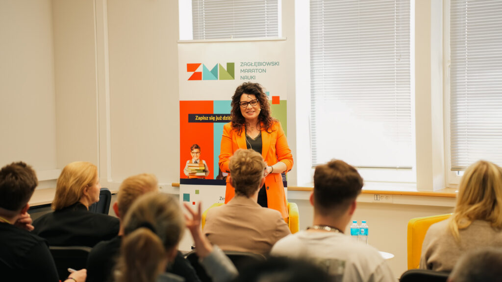 Prelegentka w pomarańczowej marynarce prowadzi spotkanie edukacyjne przed zgromadzoną publicznością; w tle widoczny roll-up Zagłębiowskiego Maratonu Nauki.