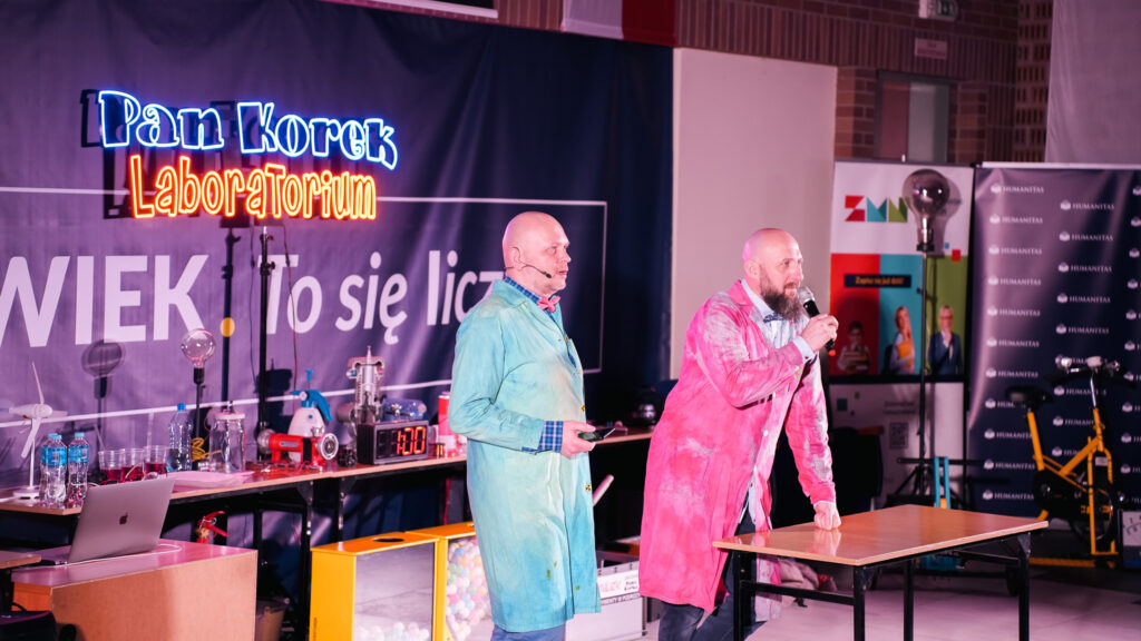 Dwóch prowadzących w kolorowych fartuchach laboratoryjnych prowadzi pokaz naukowy na scenie; jeden mówi do mikrofonu, drugi obsługuje rekwizyty, a w tle widoczny jest napis »Pan Korek Laboratorium«, hasło »Człowiek. To się liczy« oraz banery Akademii Humanitas i Zagłębiowskiego Maratonu Nauki.