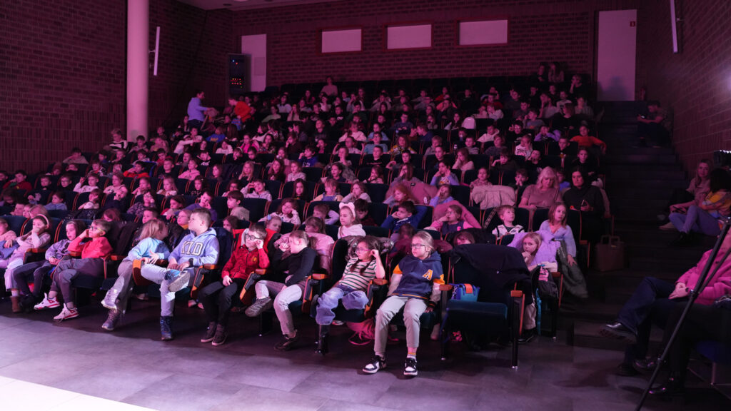 Widownia wypełniona dziećmi siedzącymi w fotelach sali audytoryjnej, oświetlonej różowym światłem, podczas wydarzenia edukacyjno-naukowego.