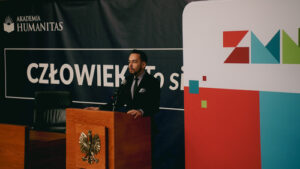 Prelegent stoi przy mównicy i przemawia do publiczności; w tle baner „Człowiek. To się liczy” oraz logo Zagłębiowskiego Maratonu Nauki.